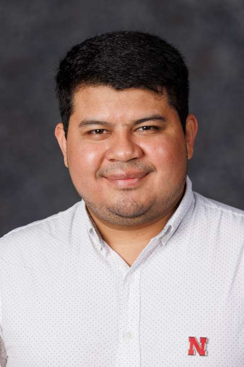 Dr. Teddy Garcia-Aroca wearing a white button down shirt with a red "N" pin on the chest, in front of a dark grey background.