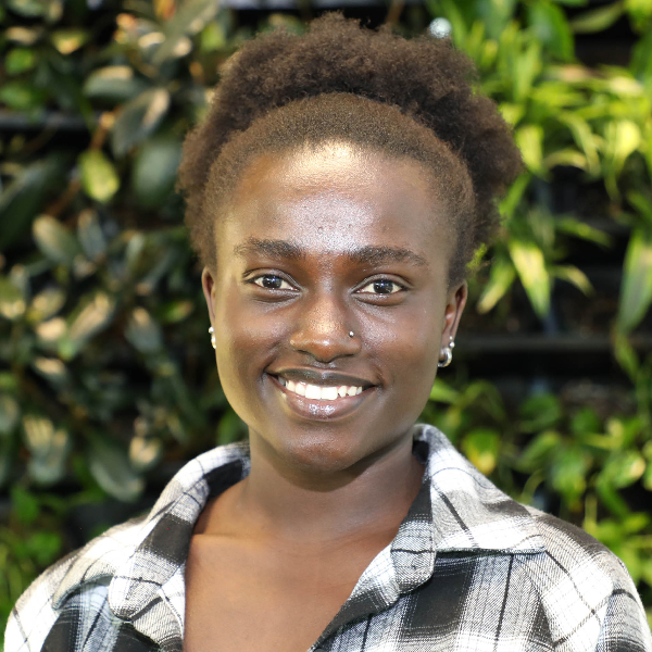 Norah Tumusiime stands in front of a backgroup of greenery in a black, white, and gray plaid shirt. 