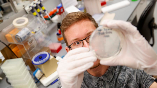 A student in white rubber gloves holds up a petri dish with black writing.