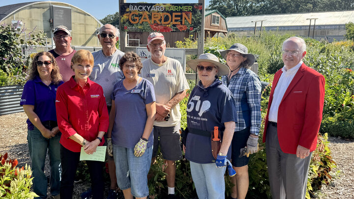 Nine people, seven in gardening clothes and two in business dress, pose for a photo in front of a garden.